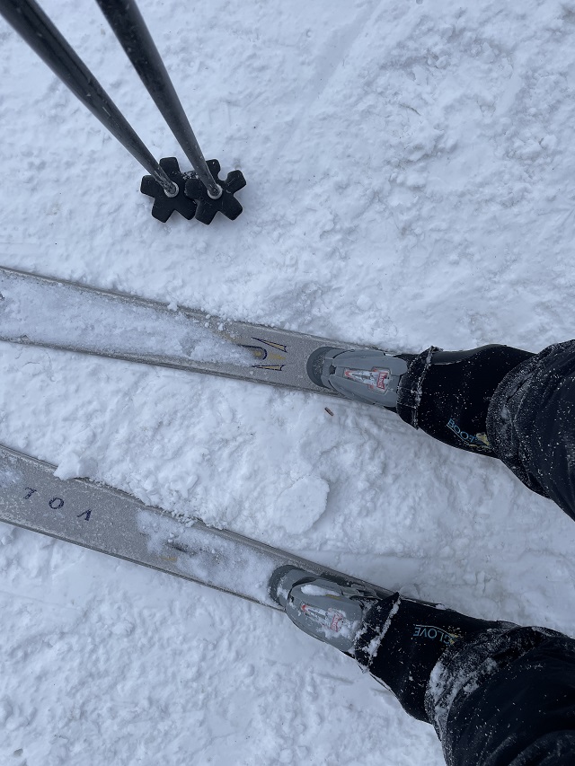 Looking down at a skier's legs standing on the snow, boots clicked into stainless steel colored skis and the bottoms of two ski poles visible in the upper part of the frame.