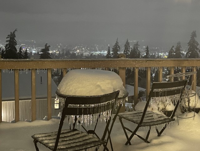 Two black metal chairs on either side of a small table. The chairs and table are sitting in maybe half a foot of snow, with a similar amount piled on the table. The chairs have icicles hanging off the slats on the backs of the chairs.