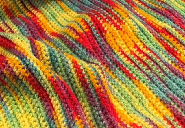 Closeup of a crocheted afghan made from variegated yarn in yellow, yellow orange, red, warm purple, turquoise, teal, and pale green. The piece is not laying quite flat but has some ripples and folds.