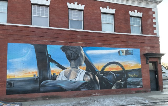 Mural painted on the side of a red brick building depicting a woman wearing a white tank top behind the wheel of a car. The woman has her hair up in a bun and is viewed from behind. An indistinct cityscape can be seen through the windows of the car.