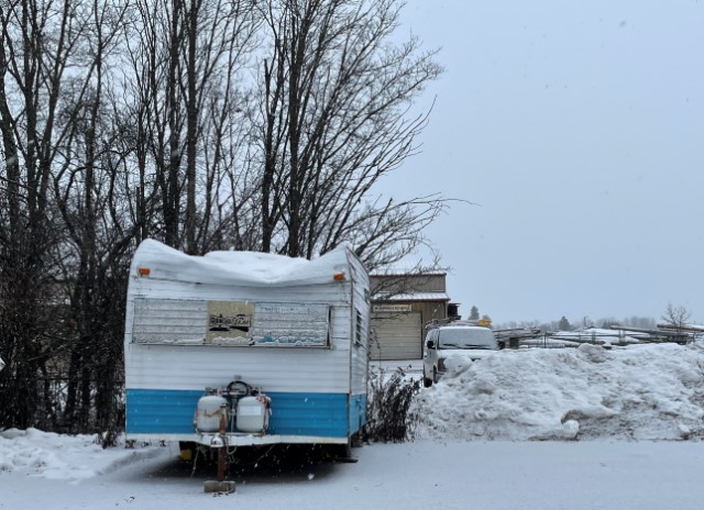 Snowy parking lot with a small camper trailer backed in to one of the spaces.