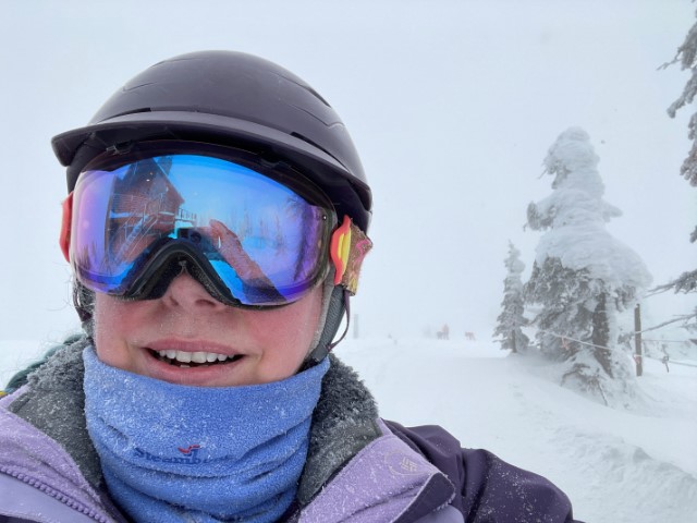 Me, a white woman, suited up for skiing in helmet, goggles, neck gaiter, and winter coat, standing at the top of a ski run with almost whiteout conditions in the background. 