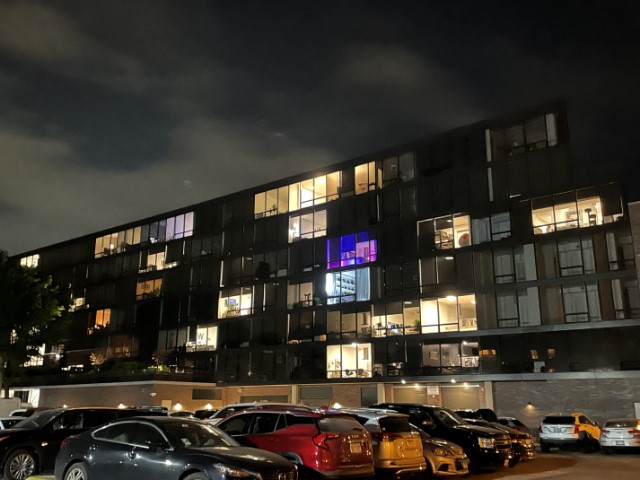 City scene at night. Parked cars in a lot in the foreground. Seven-story apartment building in the background with some apartment windows lit and some dark. One window is lit in blue and pinks and purples.