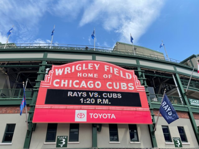 Red marquee with white lettering reading Wrigley Field, Home of the Chicago Cubs. An electronic signboard build into the marquee says Rays vs. Cubs, 1:20 p.m.
