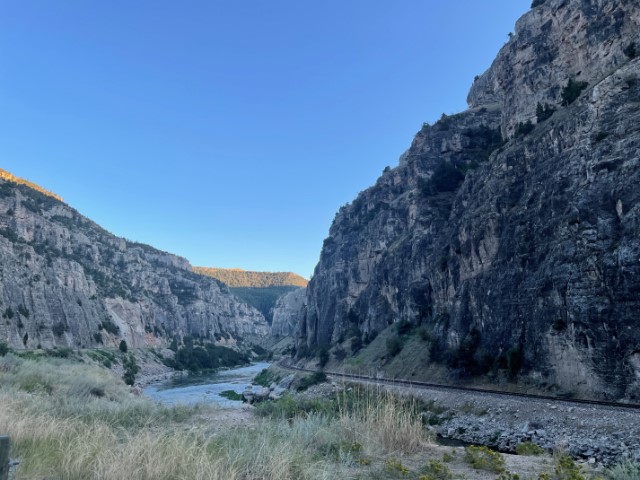 Looking down a canyon with high grey rock wall and a road, river, and train tracks at the base.