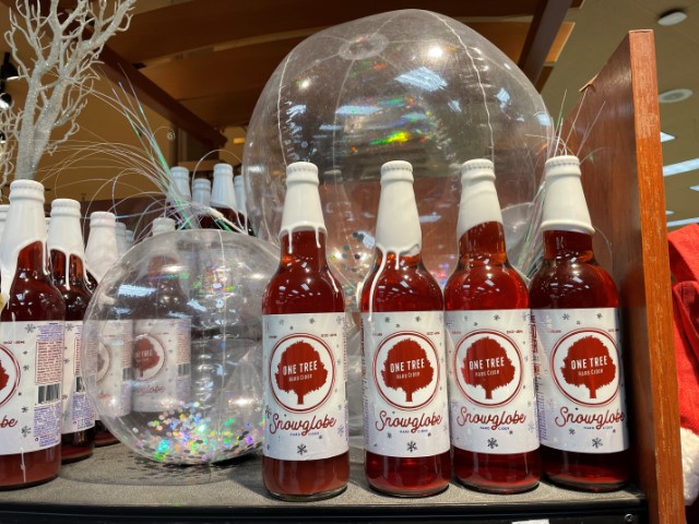 Store shelf with bottles of Snowglobe cider, which have berry red liquid and white wax seals on the caps. In between sections of bottles, there are two clear inflatable balls with holographic silver glitter inside, so when you pick up a ball and shake, it looks like a snowglobe.