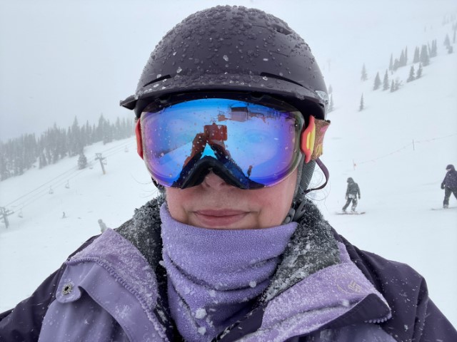 Self portrait in my ski gear, standing on a run with a lift over my shoulder on the left and two snowboarders heading down the misty slope behind me. My helmet, goggles, jacket, and neck gaiter are dotted with slushy bits of snow.
