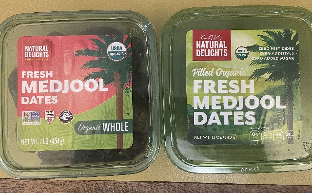 Two green translucent plastic boxes of dates sitting side by side on a yellow counter, viewed from the top. The one on the left has a red and green label; the one on the right has a mostly green label with red accents.